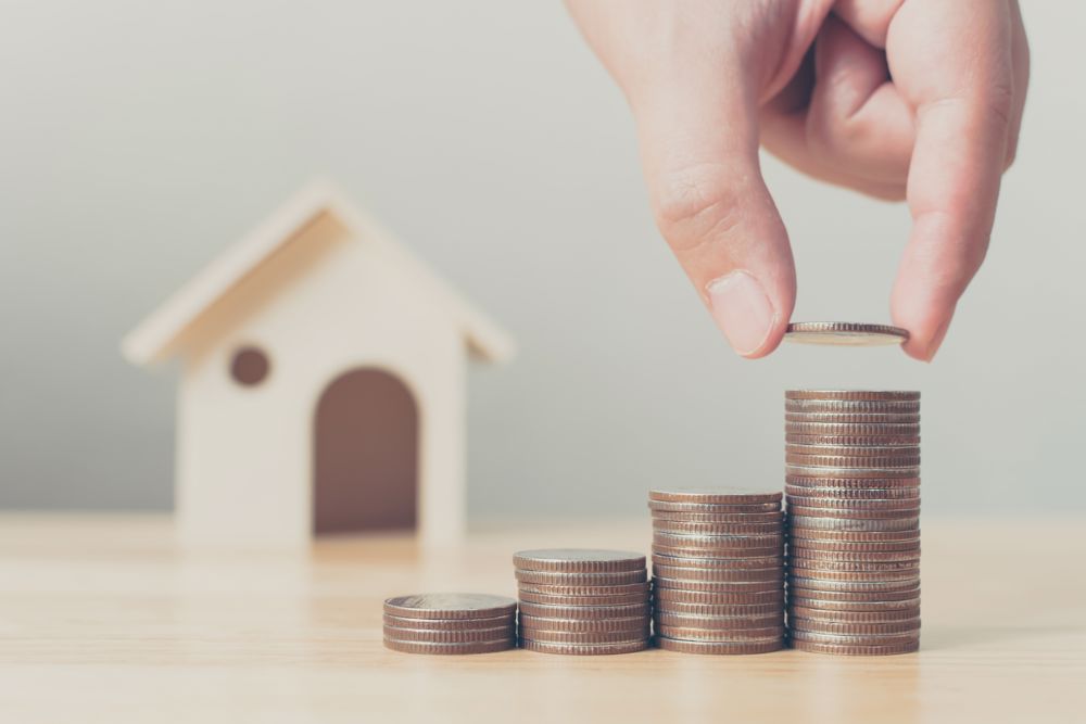 Person stacks coins in front of a miniature house.